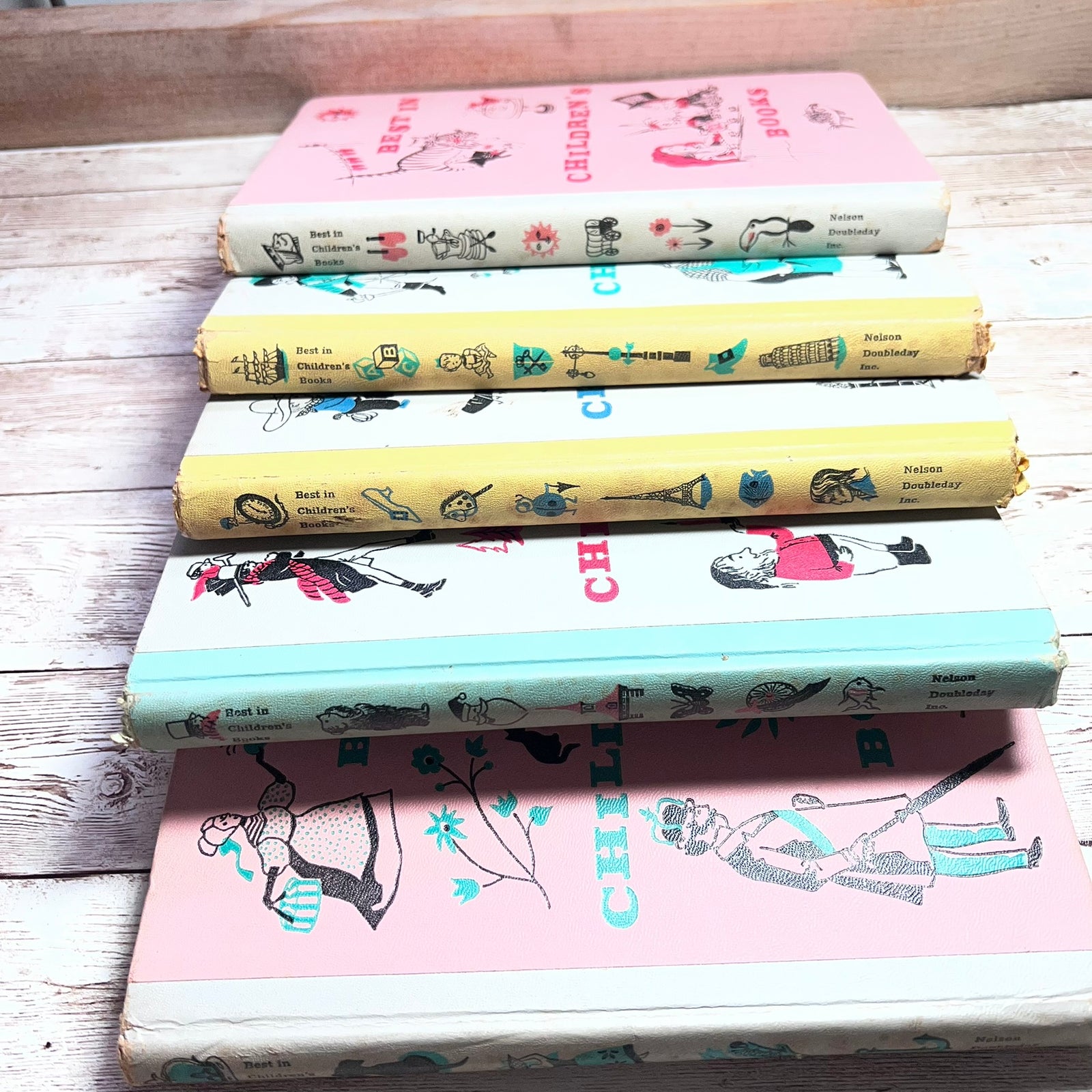 A stack of five vintage children's books with illustrated spines, arranged on a rustic whitewashed wooden surface. The spines show pastel colors including pink, yellow, blue, and white, with decorative drawings and text such as 'Children's Book' and 'Childhood'.