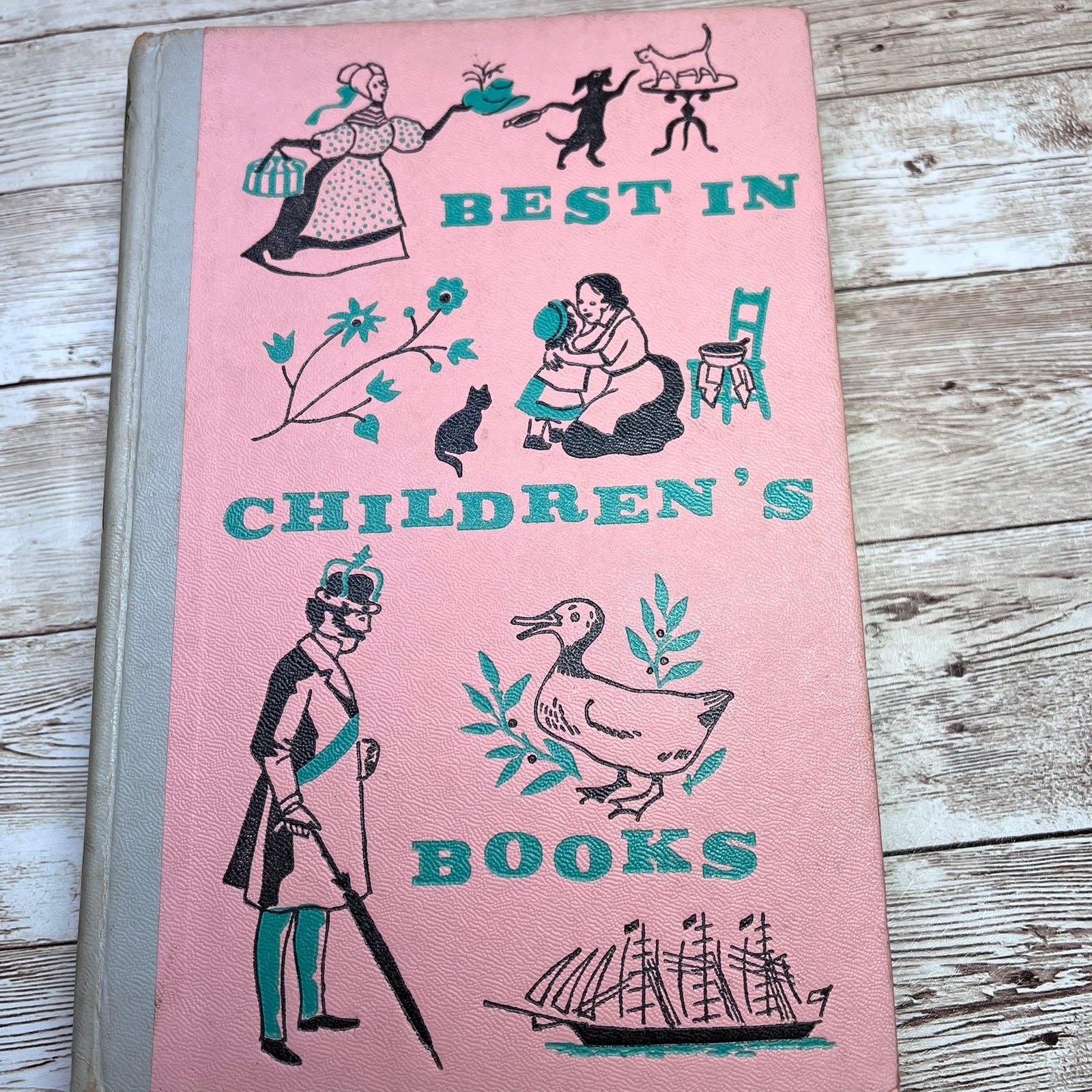 A vintage hardcover book titled 'BEST IN CHILDREN'S BOOKS' with a pink cover, illustrated with black and teal drawings of children, animals, and a duck, photographed on a rustic wooden surface.