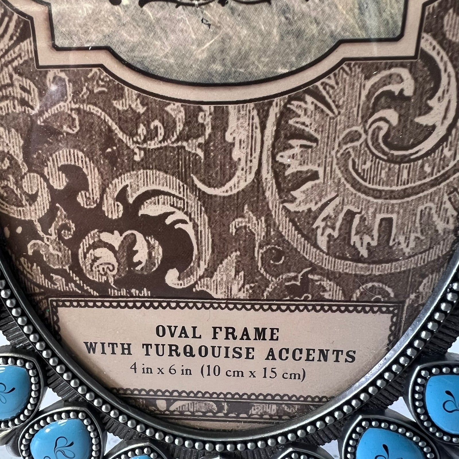 Close-up of an oval decorative picture frame with ornate brown damask-style scrollwork and turquoise blue accent stones at the bottom. Text on the frame reads 'OVAL FRAME WITH TURQUOISE ACCENTS 4 in x 6 in (10 cm x 15 cm)'.