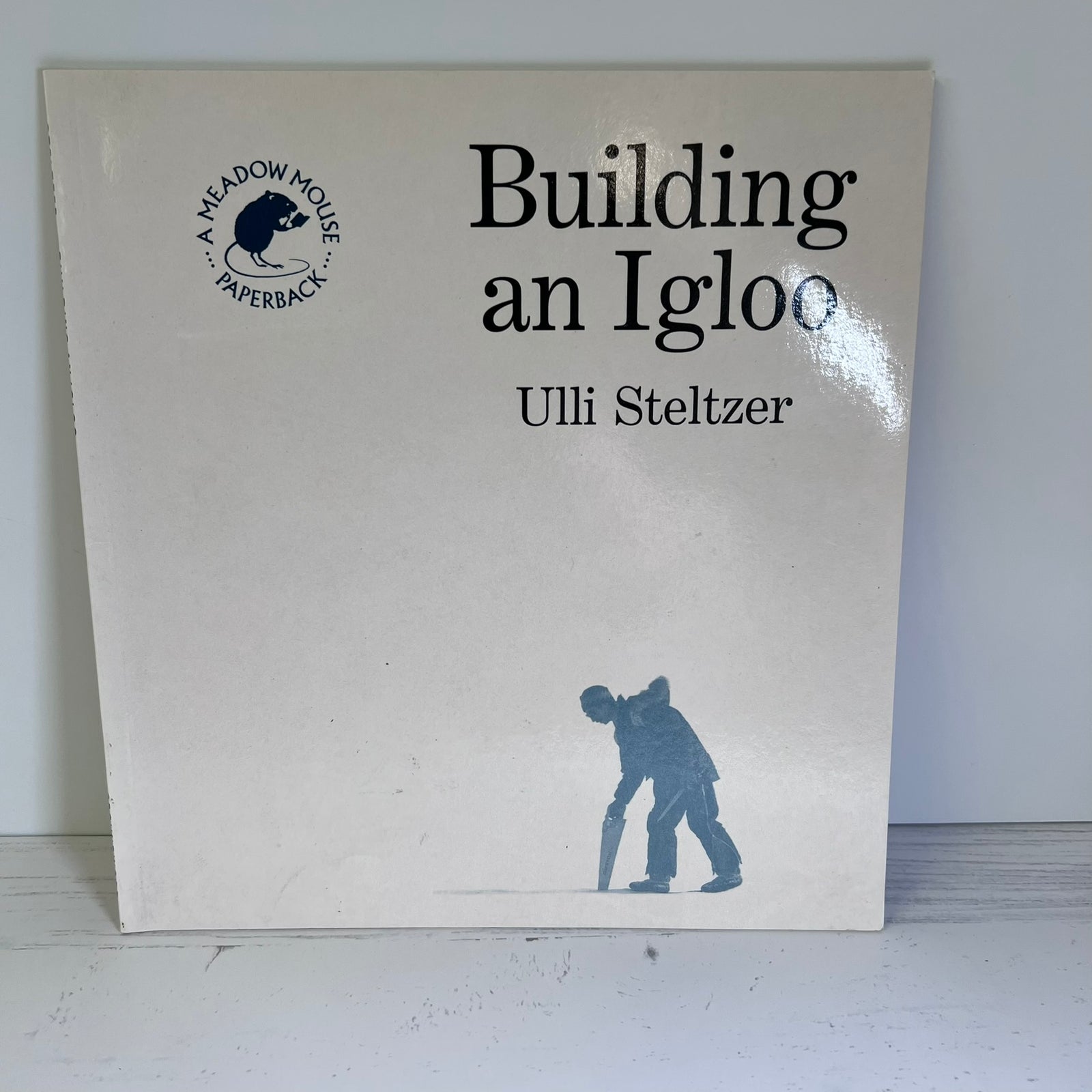 The front cover of the children's book 'Building an Igloo' by Ulli Steltzer, published by Meadow House Paperbacks. The cover is off-white with black text and a blue-gray silhouette of a child building an igloo at the bottom.