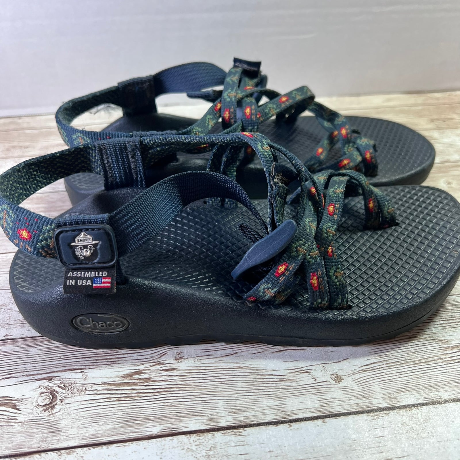 A pair of Chaco sandals with dark navy straps and footbeds, featuring a woven pattern on the front straps with small red and orange accents. The sandals are placed on a light-colored wooden surface.