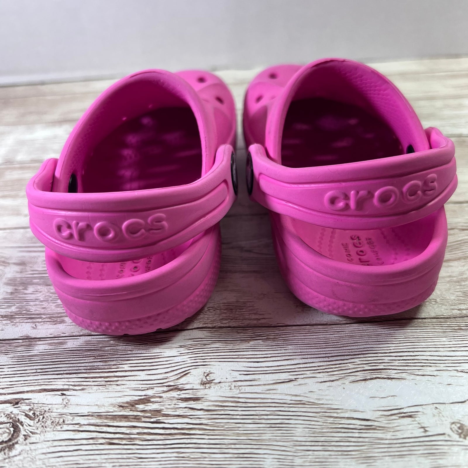 A pair of pink Crocs children's clogs shown from the back, resting on a light-colored wooden surface.
