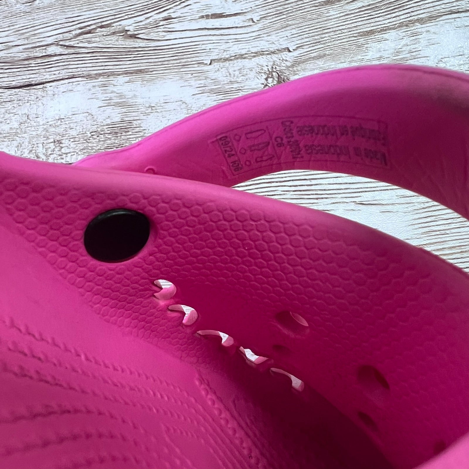 A close-up of a bright pink Croc-style clog or sandal showing the textured footbed, side profile, and inner sole with printed size and material labels, placed on a light wood surface.