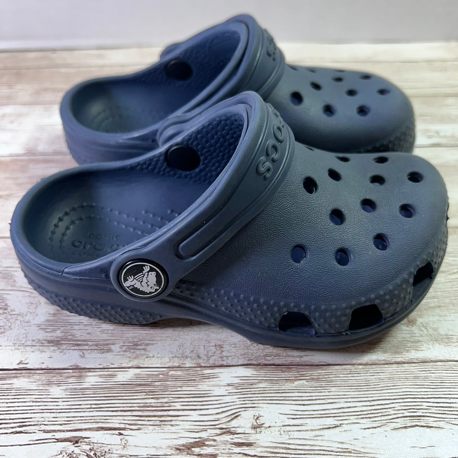 A pair of dark blue Croc-style clogs on a light wood surface, with the brand name partially visible on the strap and a Croc logo charm on the side.
