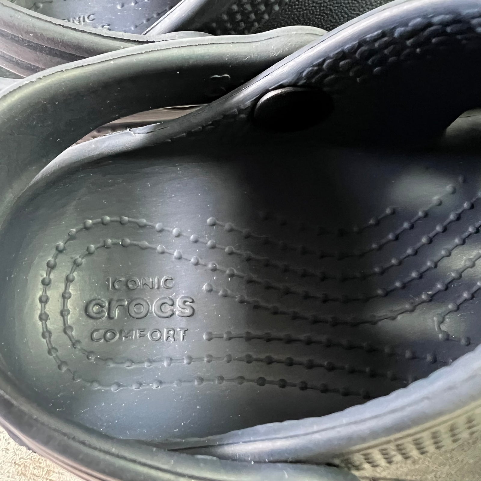 Close-up of the inside of a black Crocs clog, showing the embossed 'ICONIC CROCS COMFORT' sole and the side strap with a snap closure.