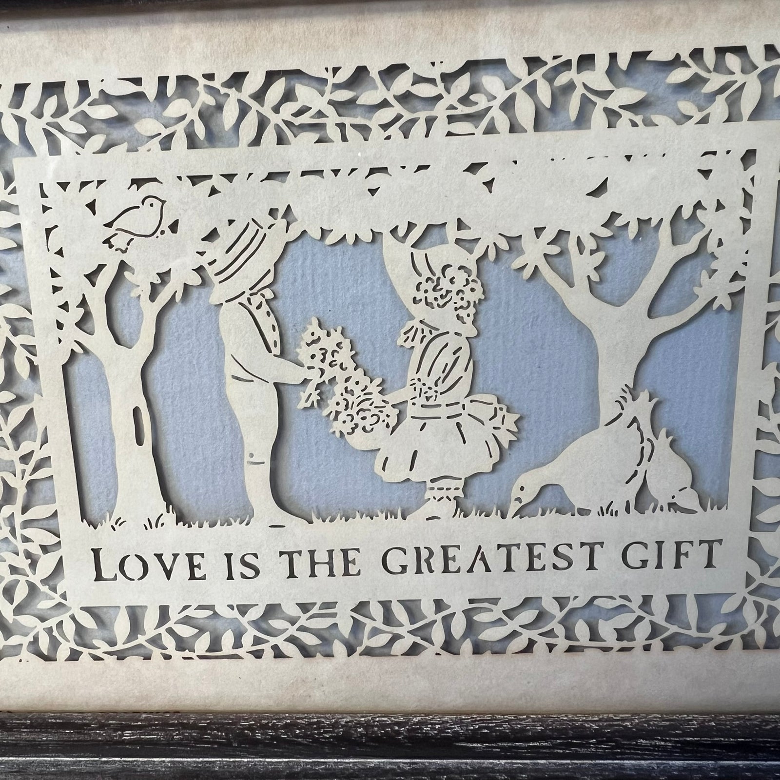 A square laser-cut wooden decorative piece with a cutout scene of a couple, trees, birds, and a chicken, with the text 'LOVE IS THE GREATEST GIFT' along the bottom, shown against a light blue backing board.