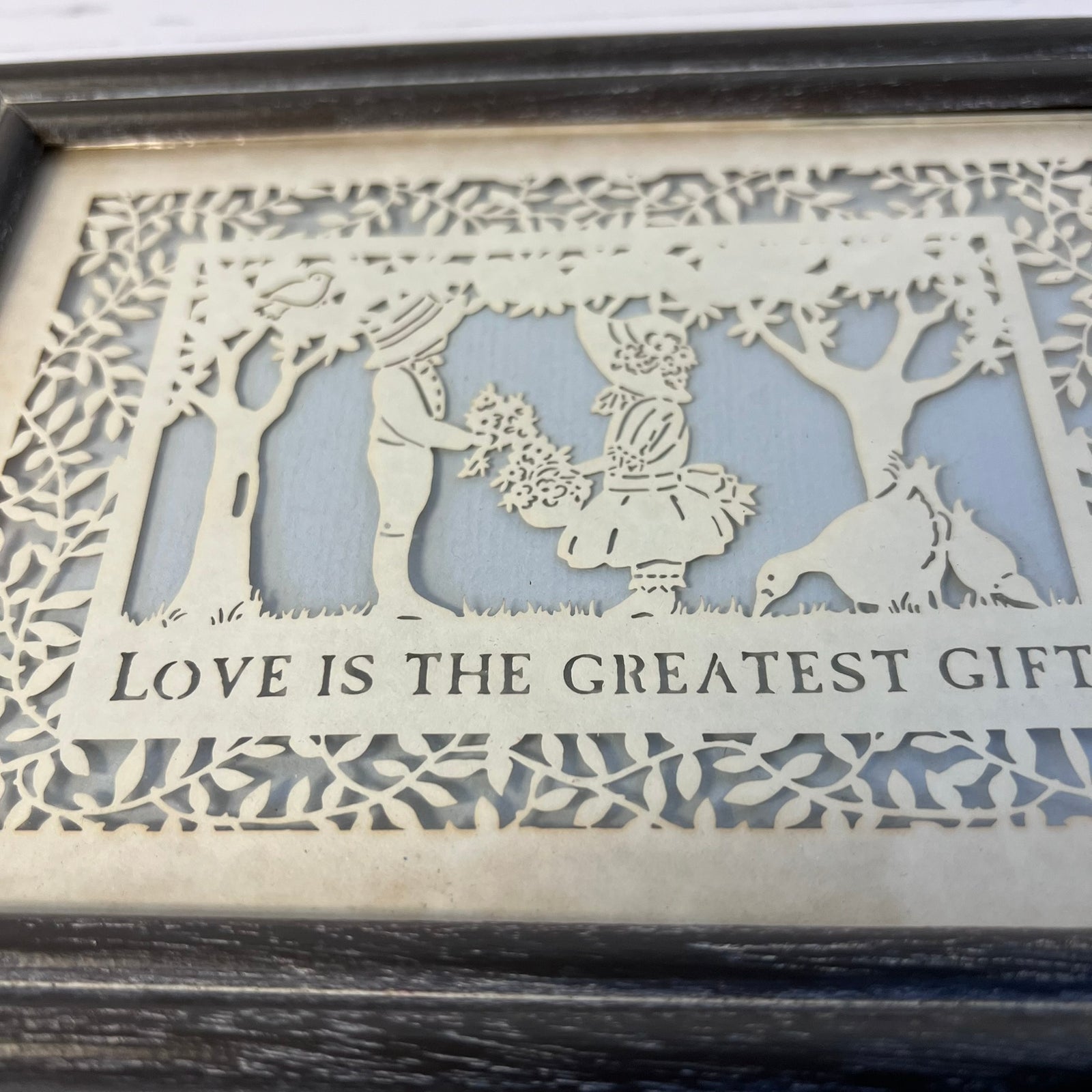 A laser-cut decorative wall art piece with a cutout scene of a couple and animals under trees, with the text 'LOVE IS THE GREATEST GIFT' across the bottom. The piece has a light beige/cream tone and is shown close-up inside a dark frame.
