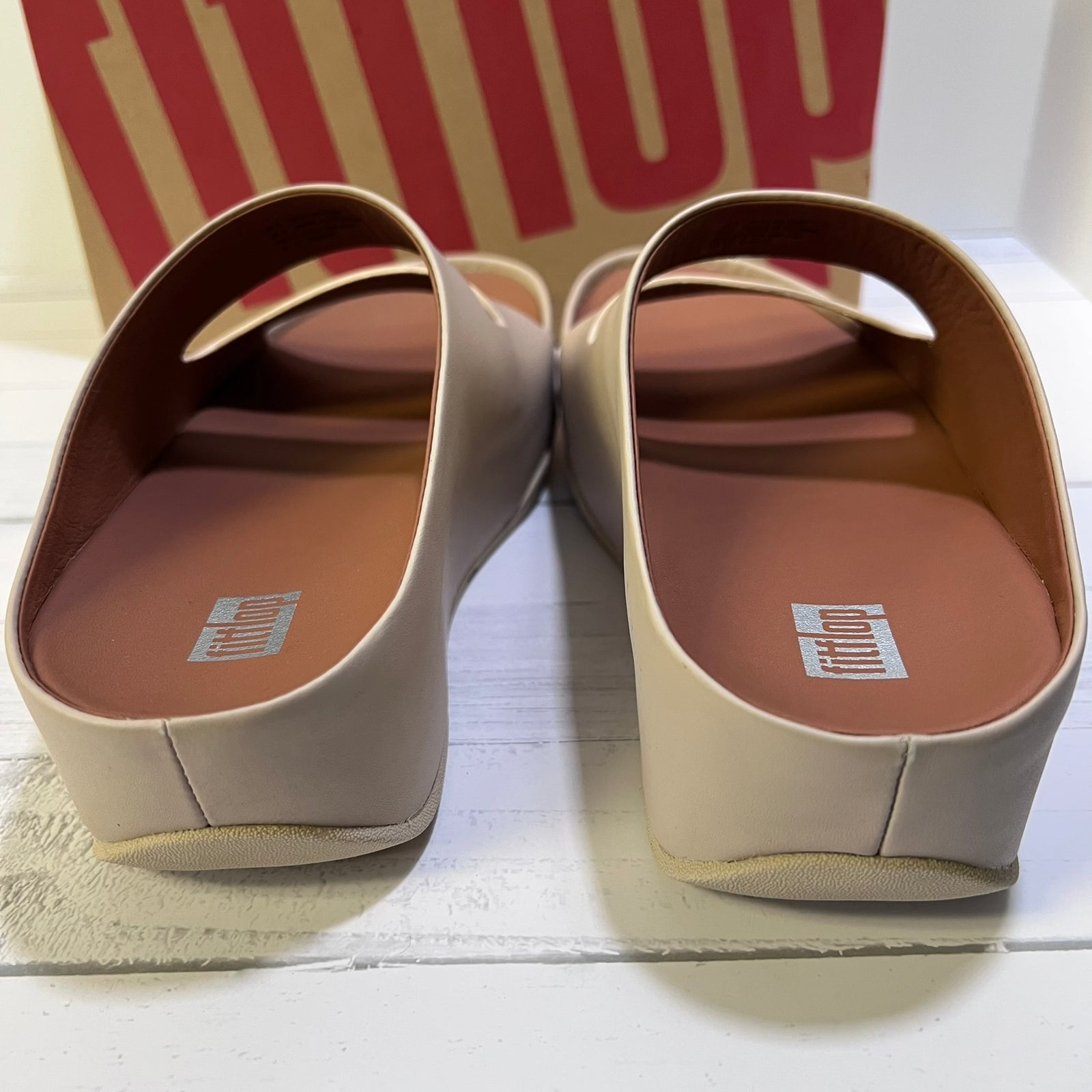 A pair of FitFlop slip-on mule sandals in a light beige color, shown from the back on a white wooden surface with the FitFlop brand box in the background.