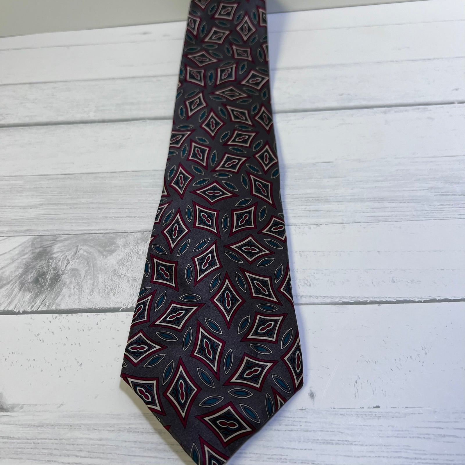 A men's necktie with a dark navy blue base and a repeating geometric diamond pattern in red, white, and teal, laid flat on a white wood plank surface.