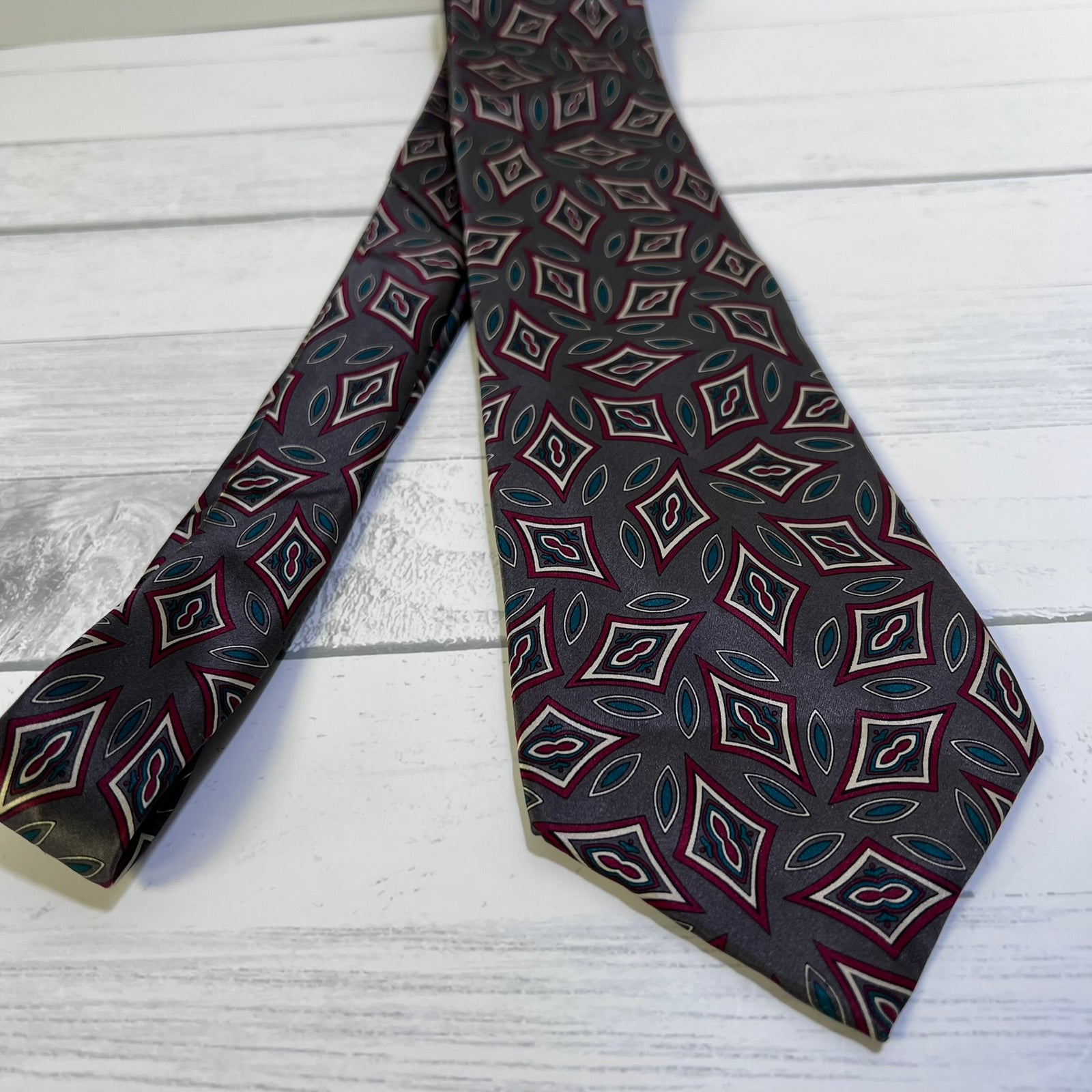 A patterned necktie with a dark gray base and a repeating geometric design in burgundy, cream, and teal, laid flat on a white wood plank surface.