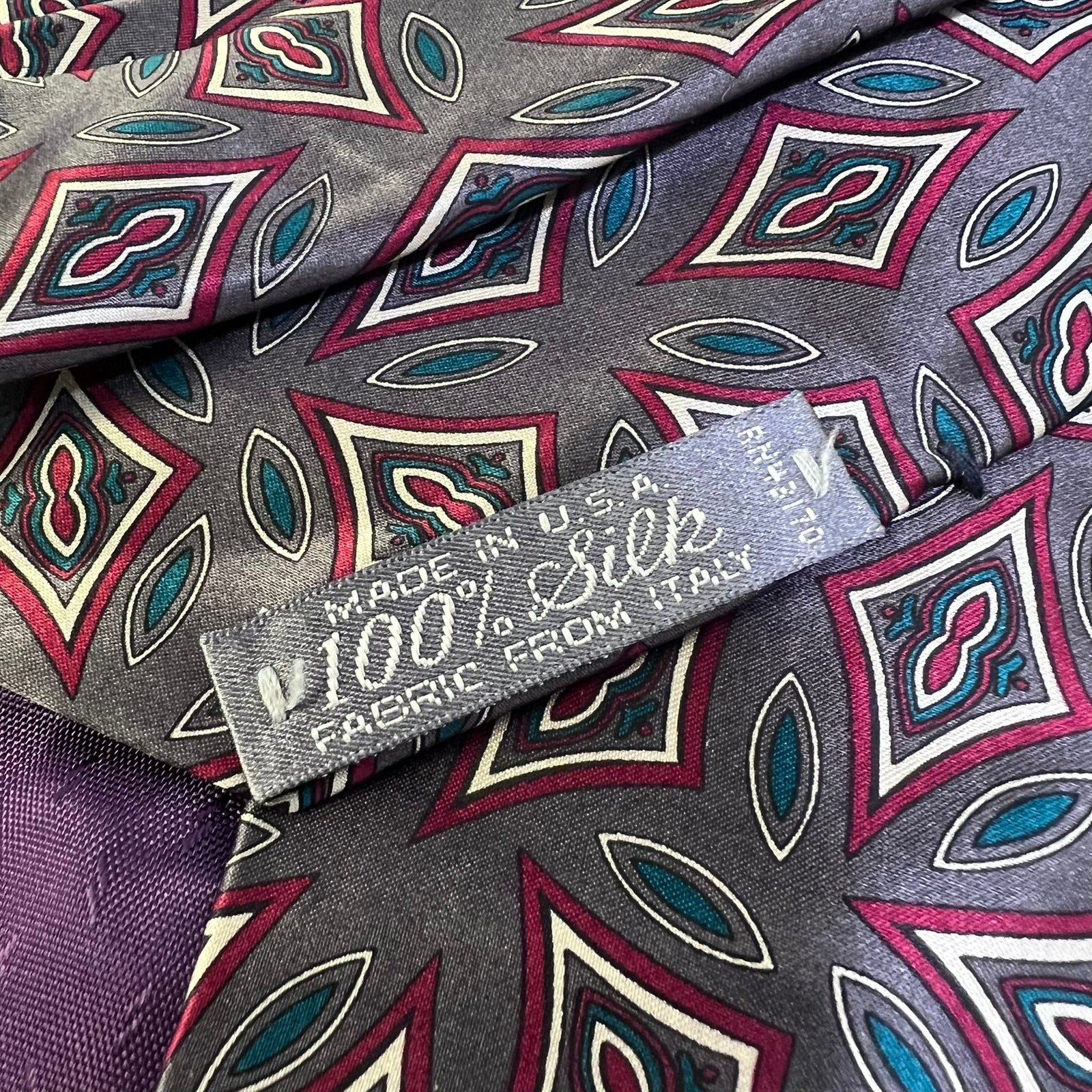 A close-up of a patterned silk fabric with a geometric design in gray, burgundy, teal, and cream, featuring a woven label that reads '100% Silk' and 'MADE IN U.S.