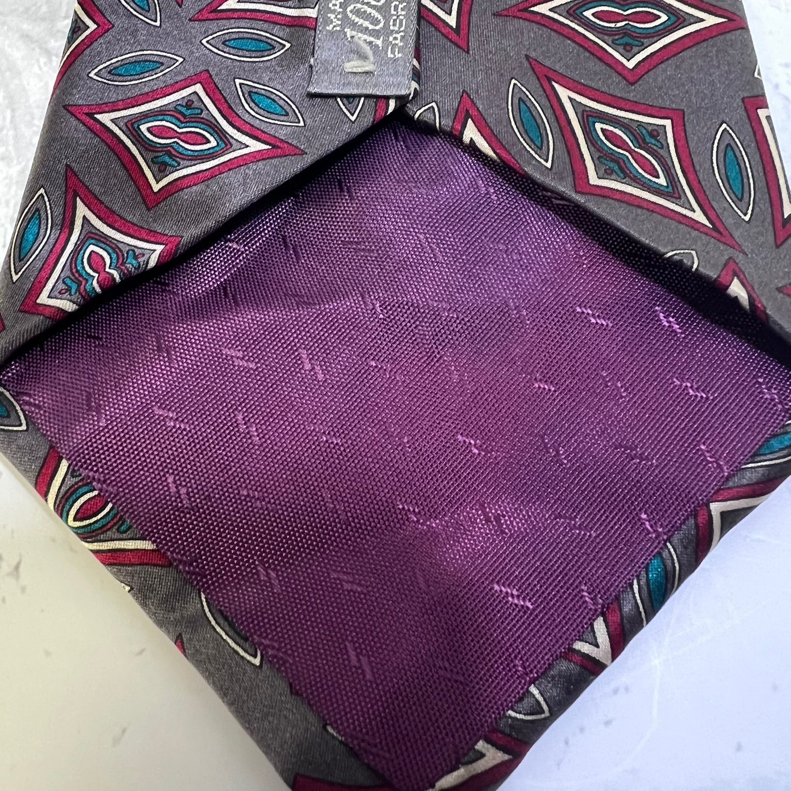 A folded men's pocket square with a purple satin-like center and a gray patterned border featuring red, teal, and white geometric motifs. A small fabric tag is visible at the top.
