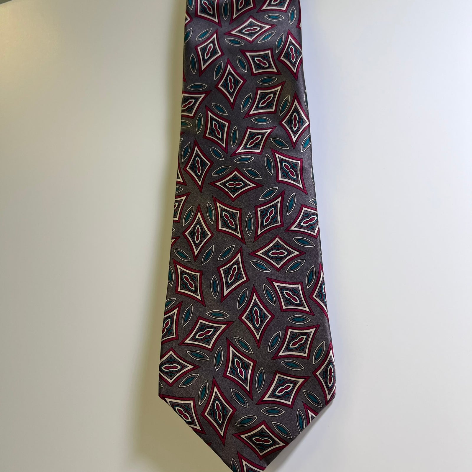A men's necktie with a repeating geometric diamond pattern in dark gray, burgundy red, white, and teal blue, hanging against a plain off-white wall.