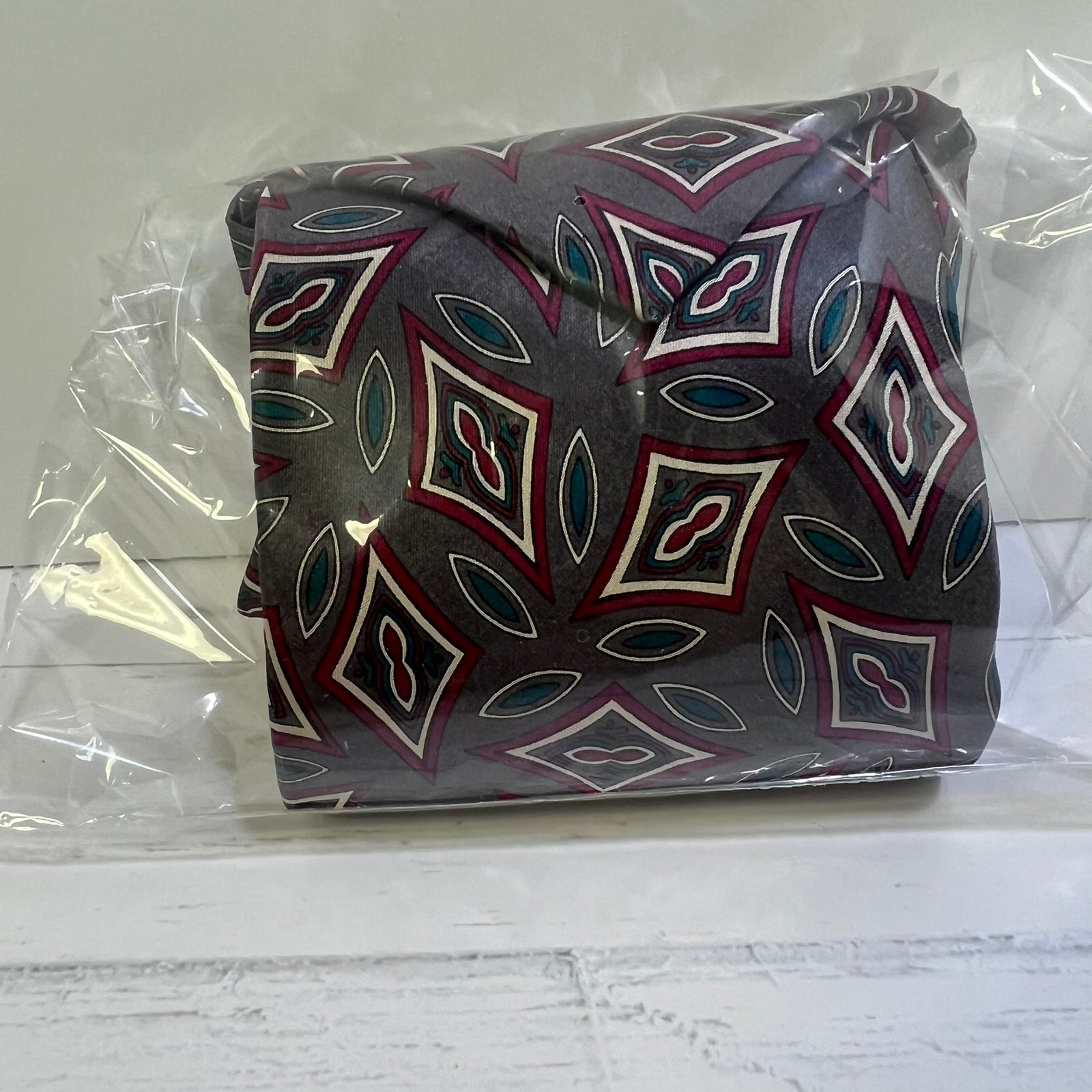 A folded patterned necktie inside a clear plastic bag, resting on a white wooden surface against a light gray background.