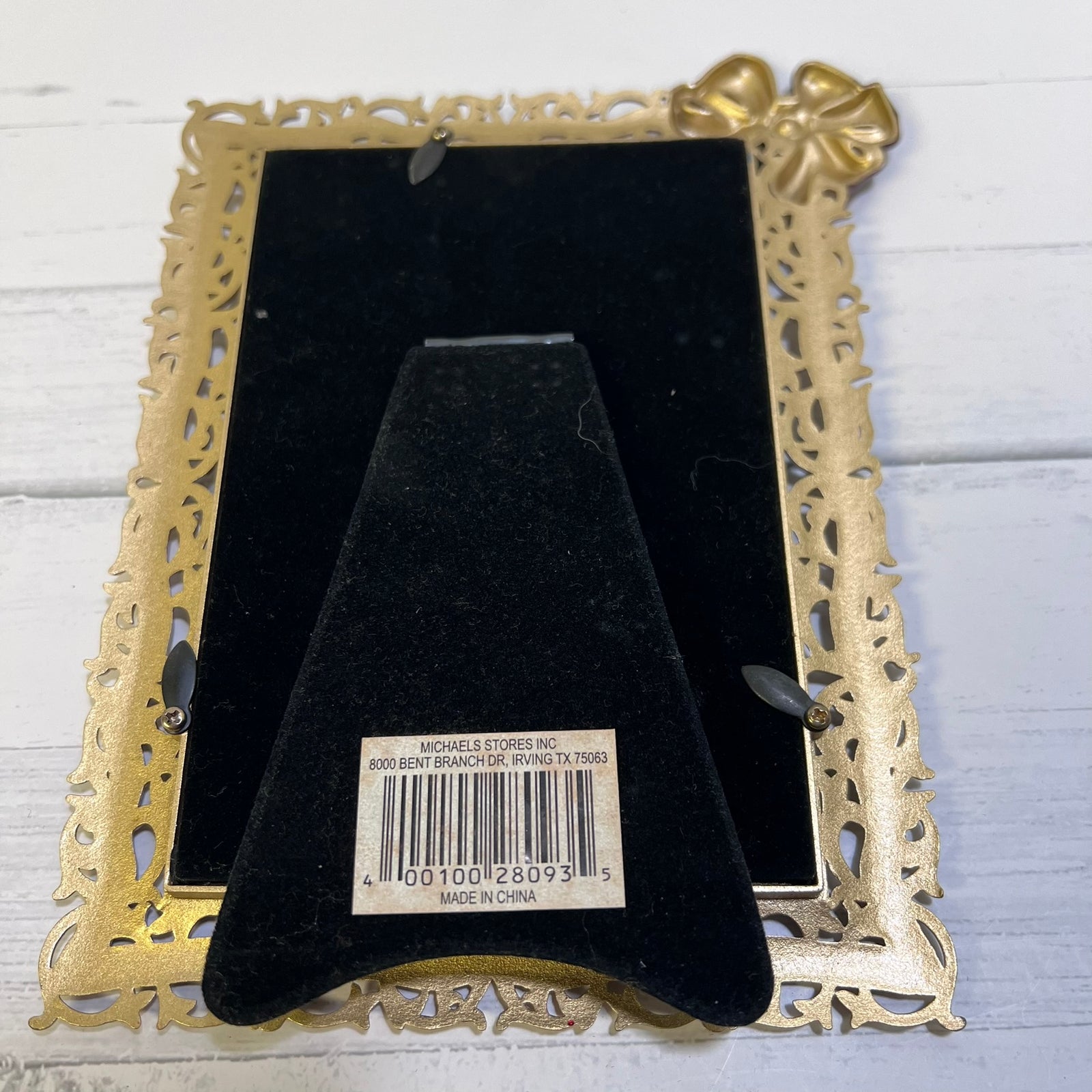 A vintage-style rectangular picture frame with an ornate filigree gold-tone border and a matching decorative corner accent, shown from the back on a white wood surface. A black velvet insert and a barcode label are visible in the center.