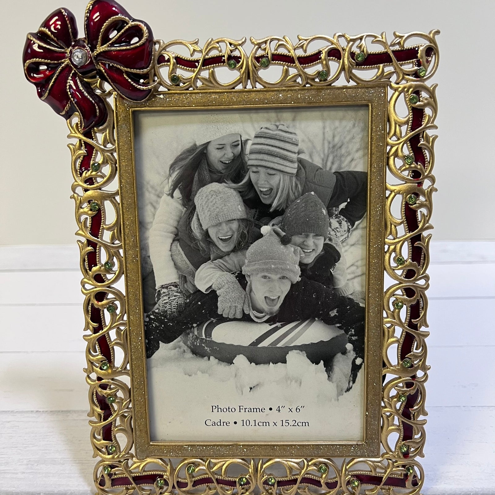 An ornate gold picture frame with red ribbon accents and a matching red bow on the top left, holding a black-and-white photo of children in snow. The frame is photographed against a light background.
