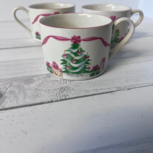 Three ceramic mugs with Christmas tree and bow designs on a white wooden surface.