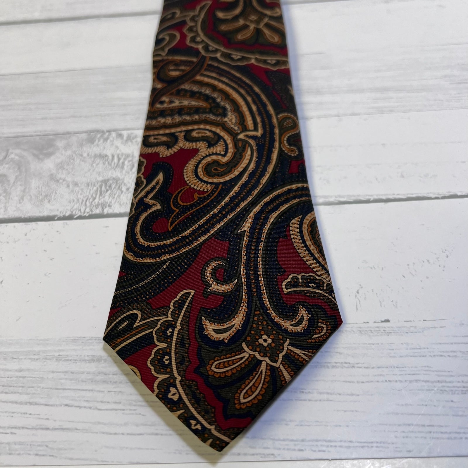 A necktie with an intricate paisley pattern in deep red, black, and gold, laid flat on a white-washed wooden surface.