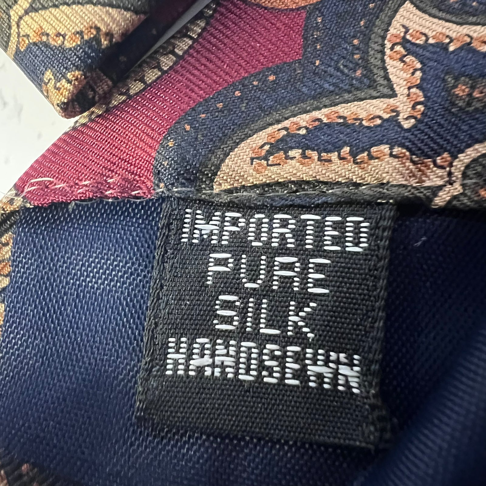 Close-up of a black woven label reading 'IMPORTED PURE SILK HANDSEWN' attached to a patterned silk garment in navy, burgundy, and tan.