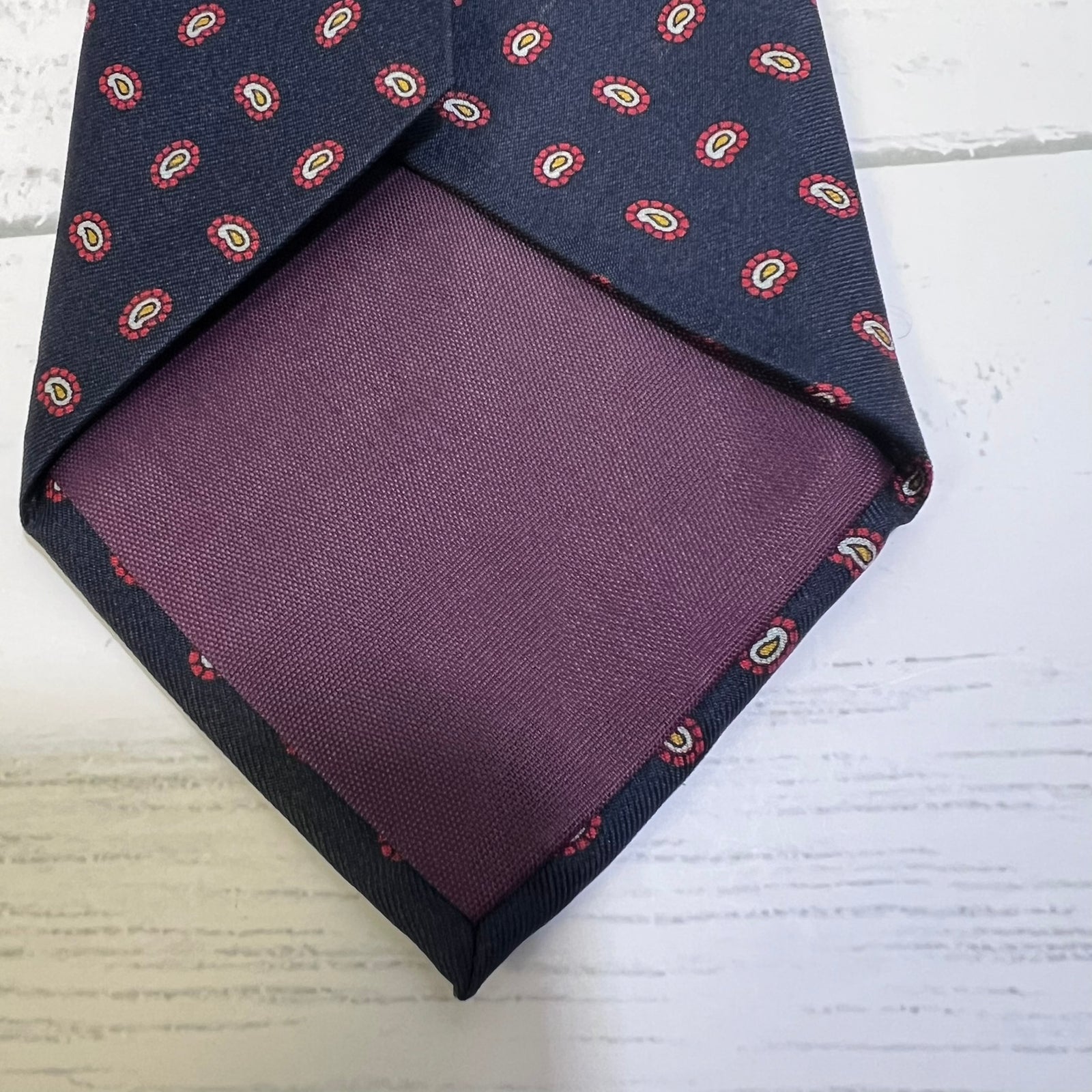 A folded necktie with a navy blue base, featuring a small red, yellow, and white paisley pattern along the outer edge and a solid deep purple inner lining, displayed on a white wood-textured background.