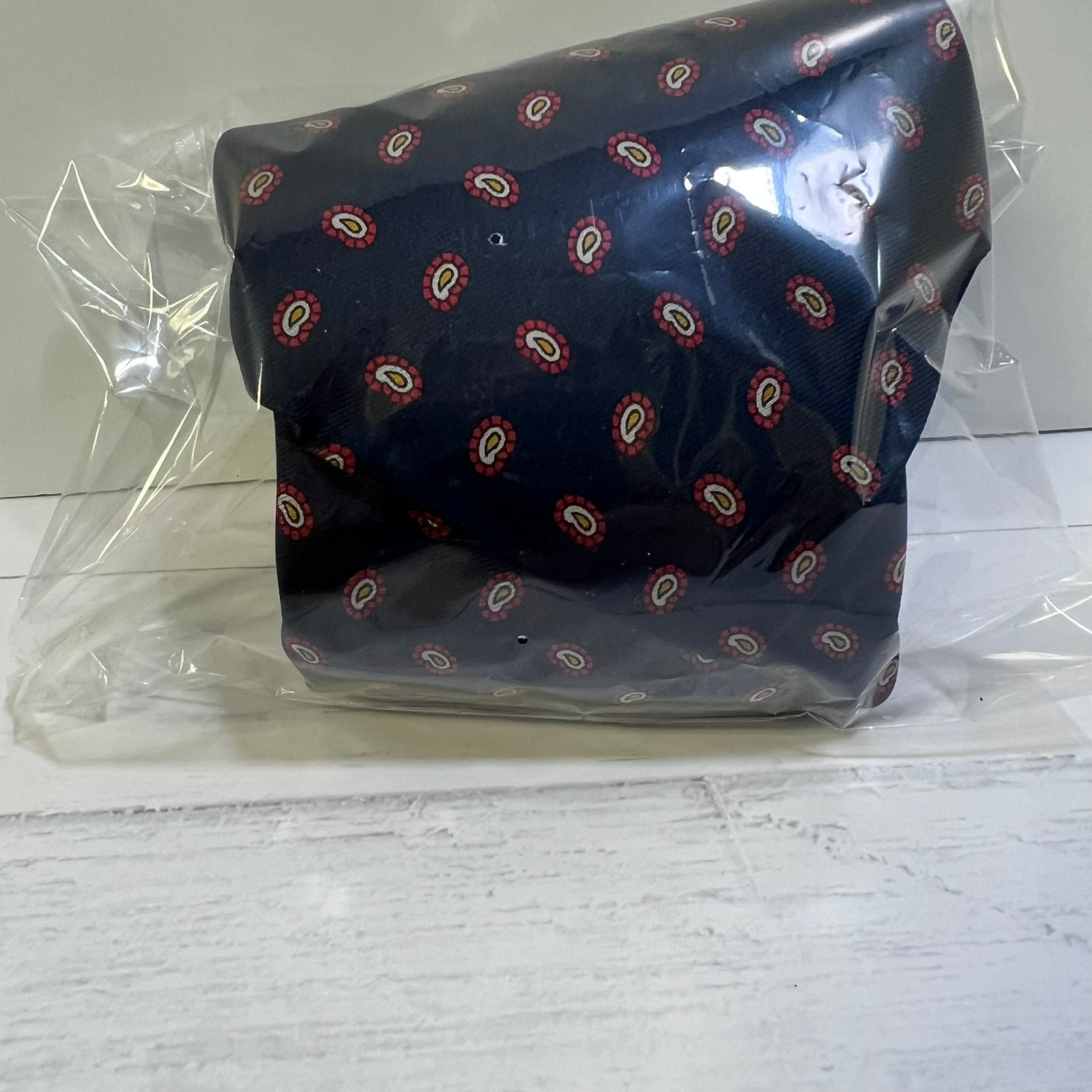 A dark navy blue fabric item, likely a pocket square or small fabric accessory, featuring a repeating red, white, and beige paisley pattern, wrapped in clear plastic packaging on a white wooden surface.