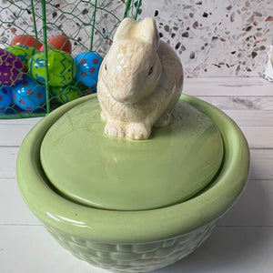 Green ceramic bowl with a rabbit-shaped lid on a wooden surface, colorful eggs in the background.