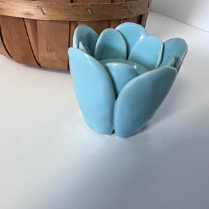 Light blue ceramic flower-shaped candle holder on a white surface with a wooden basket in the background.
