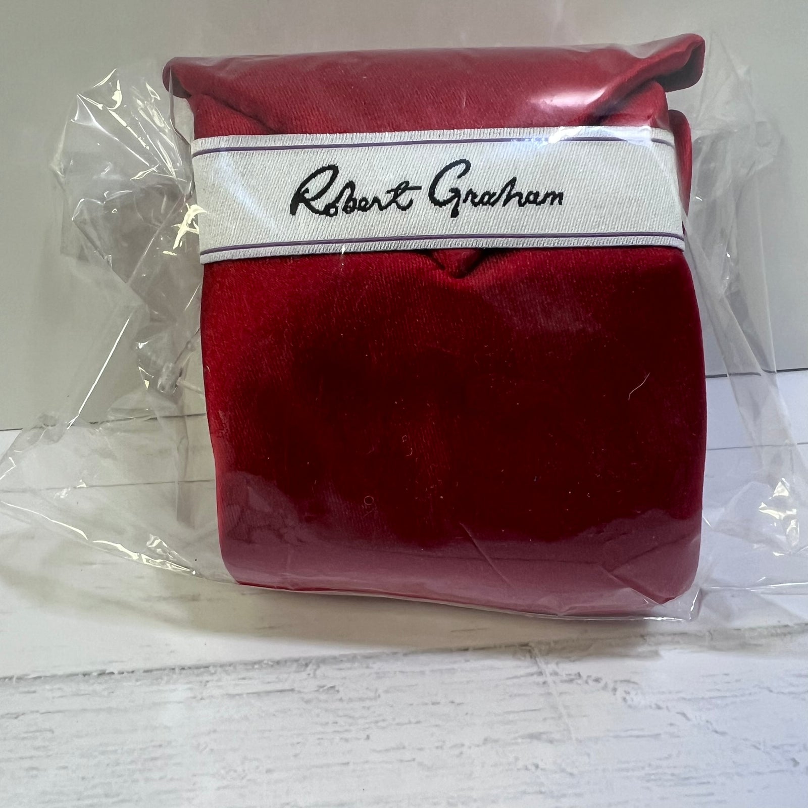 A red velvet pouch or case with a white band featuring the black embroidered script 'Robert Graham', shown inside clear plastic wrapping on a white surface.