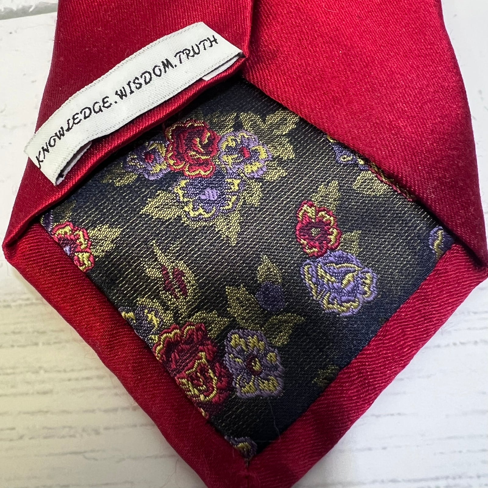 A close-up of a necktie with a deep red satin border and a dark navy floral brocade center. A white fabric label is sewn onto the collar, printed with the words 'KNOWLEDGE.