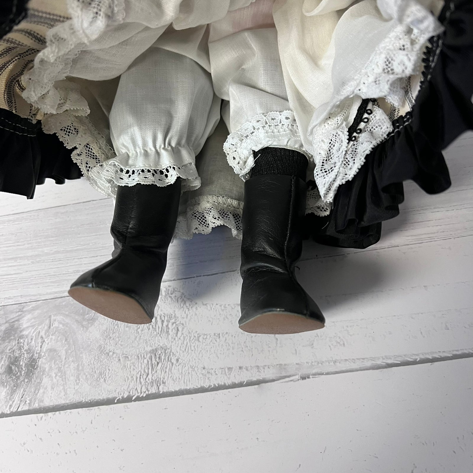 Black leather doll boots with beige soles, shown on a white ruffled dress with lace trim, photographed from a top-down angle on a white wooden surface.