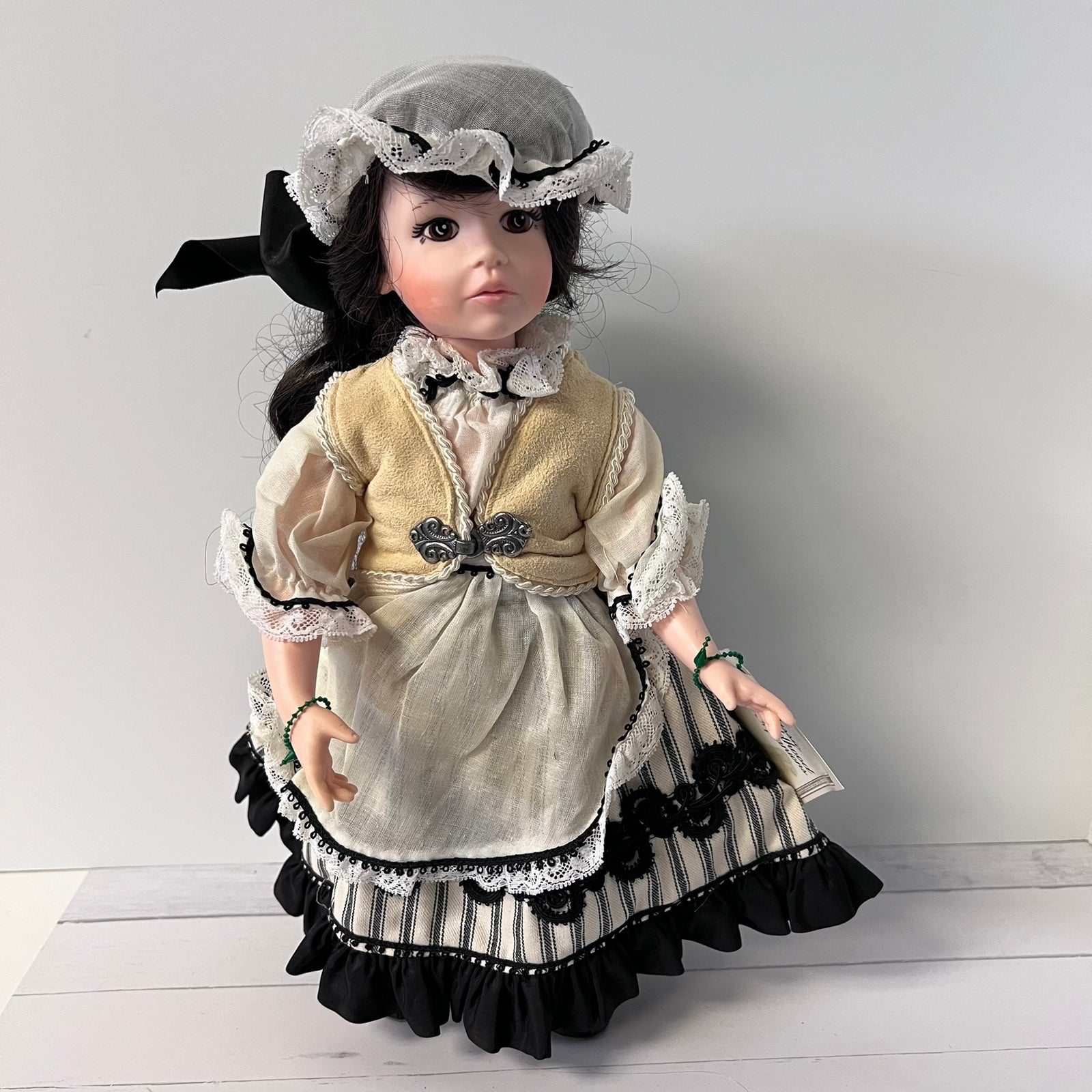 A vintage-style porcelain doll with dark hair wearing a beige and black dress with white lace trim, a matching bonnet, and green bracelets, posed against a plain light gray background.
