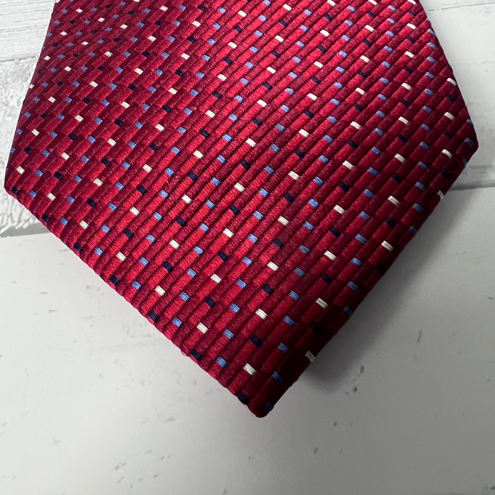 A close-up of a red necktie with a textured, woven-like pattern of small blue, white, and dark red dots, laid flat on a light gray surface.