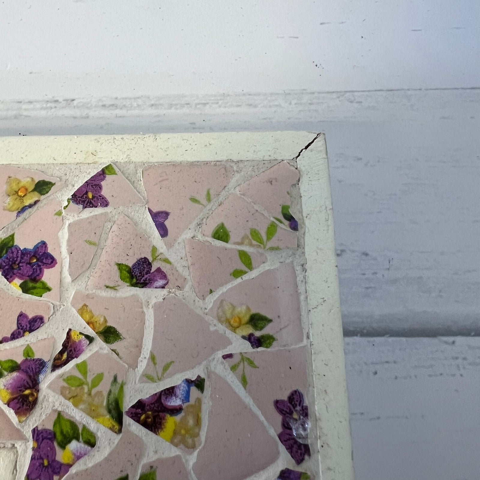 A close-up of a square mosaic tile piece with a white distressed border, featuring pink tiles and a floral pattern of purple pansies, yellow flowers, and green leaves, set against a white painted wood background.