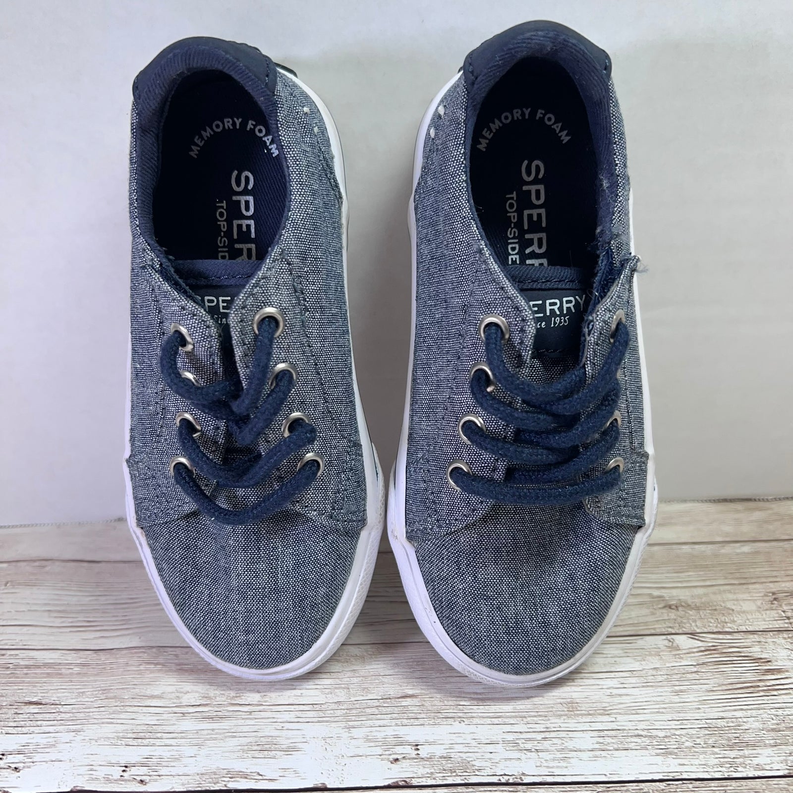 A pair of Sperry low-top canvas sneakers in heather gray with navy blue laces and white rubber soles, displayed on a rustic white wooden surface against a light gray wall.