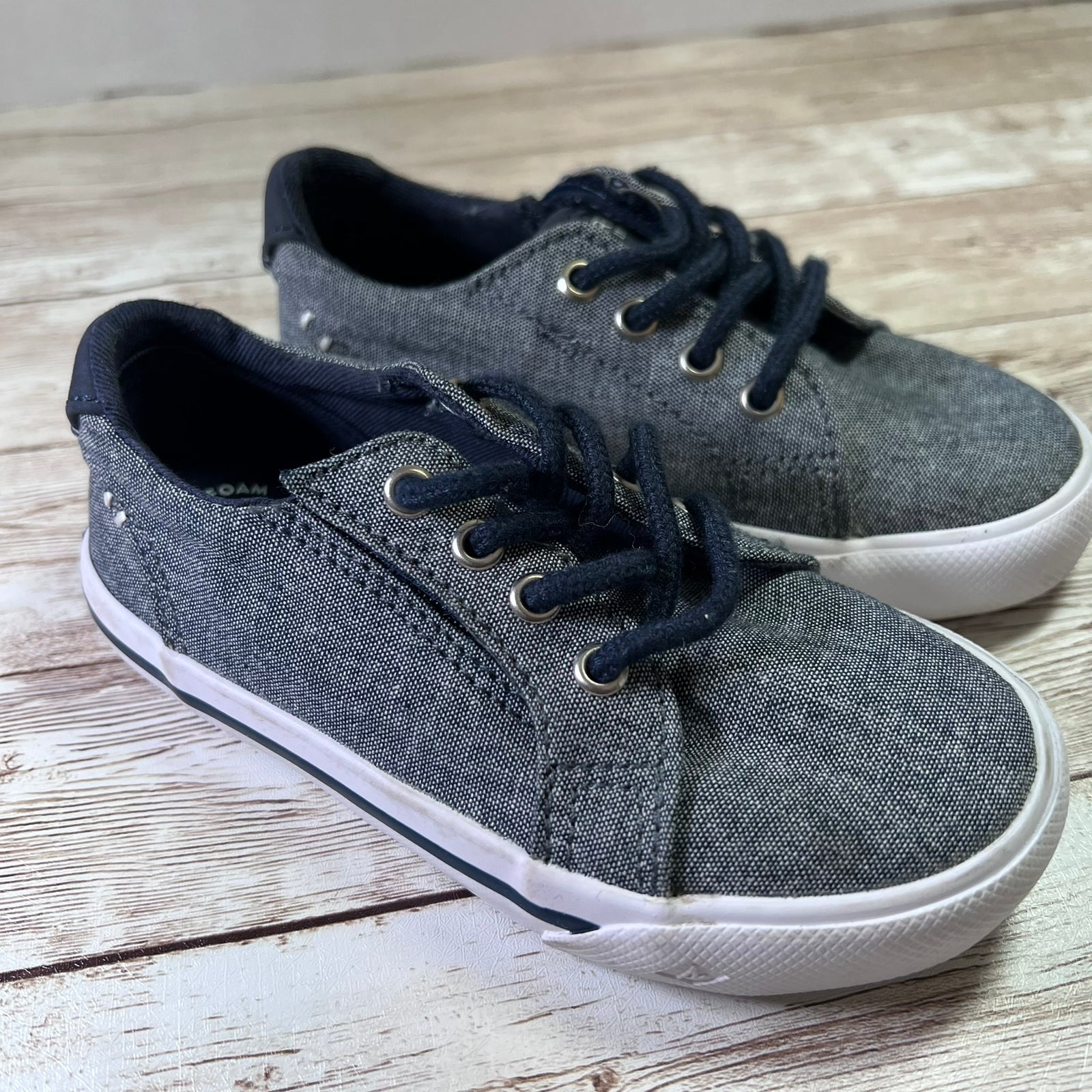 A pair of kids' low-top canvas sneakers in dark gray with navy blue laces, collar lining, and trim, and white rubber soles, displayed on a rustic light wood plank surface.