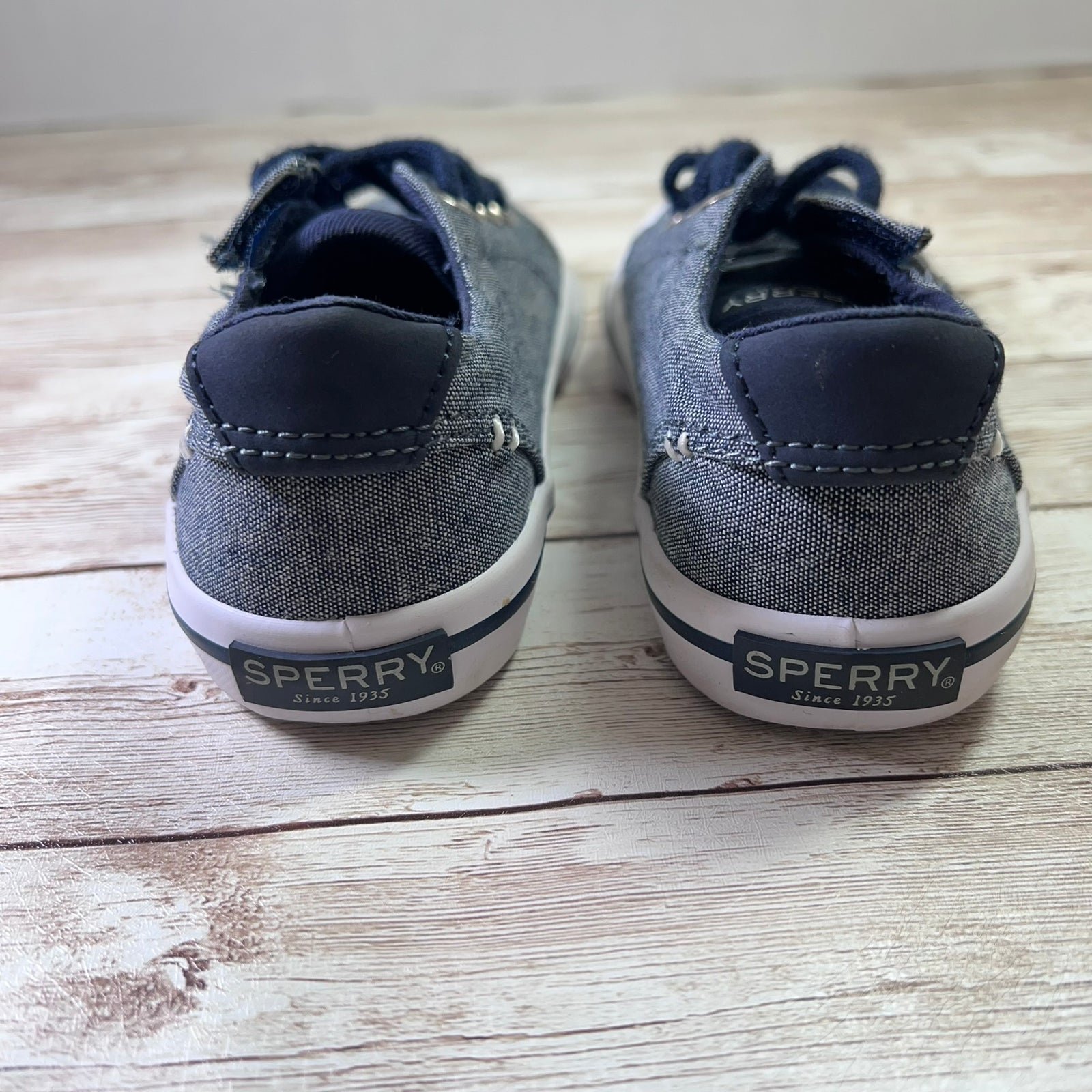 A pair of Sperry boat shoes shown from the back, featuring a dark gray denim-like canvas upper, navy blue laces and heel tabs, and a white rubber sole with a navy heel logo.