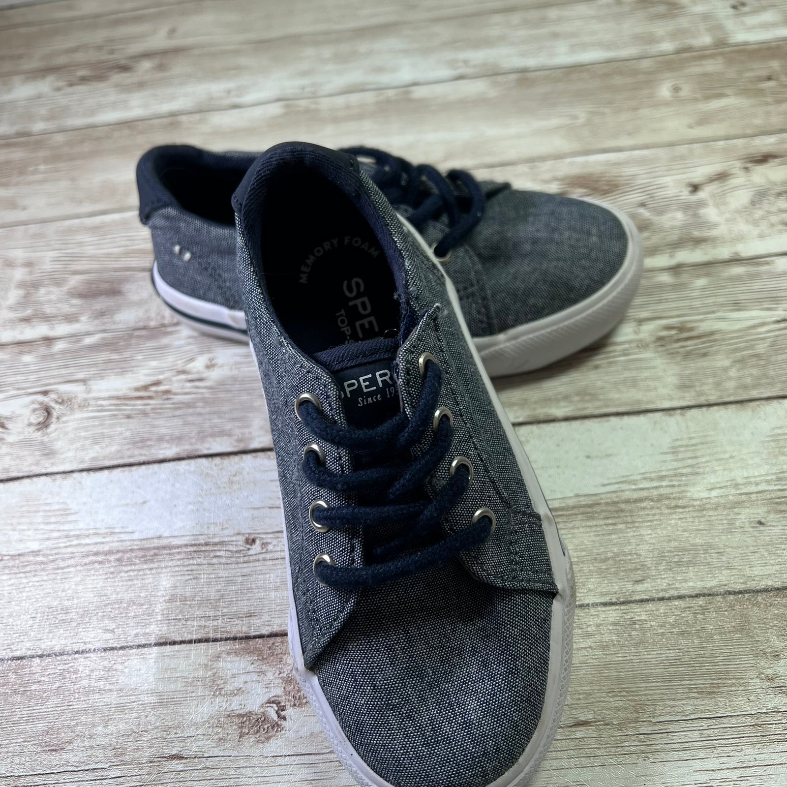 A pair of gray canvas low-top sneakers with navy blue laces and white rubber soles, displayed on a light-colored wooden plank floor. The brand name 'Sperry' is visible on the tongue label.