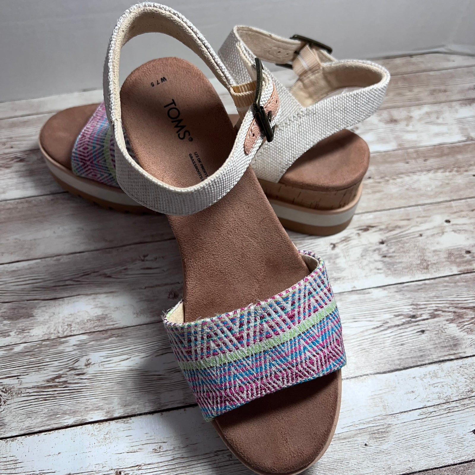 A pair of TOMS platform wedge sandals with woven multicolor straps and a light beige fabric ankle strap, displayed on a rustic white-washed wooden floor.