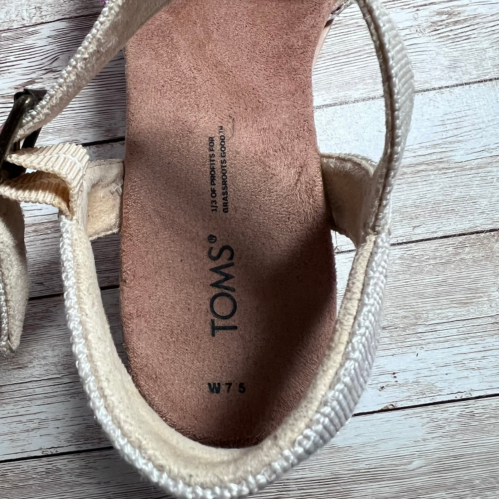A close-up of a TOMS women's sandal showing the beige woven strap, the tan suede insole with black printed branding, and the white rubber outsole, set against a light gray wood plank background.
