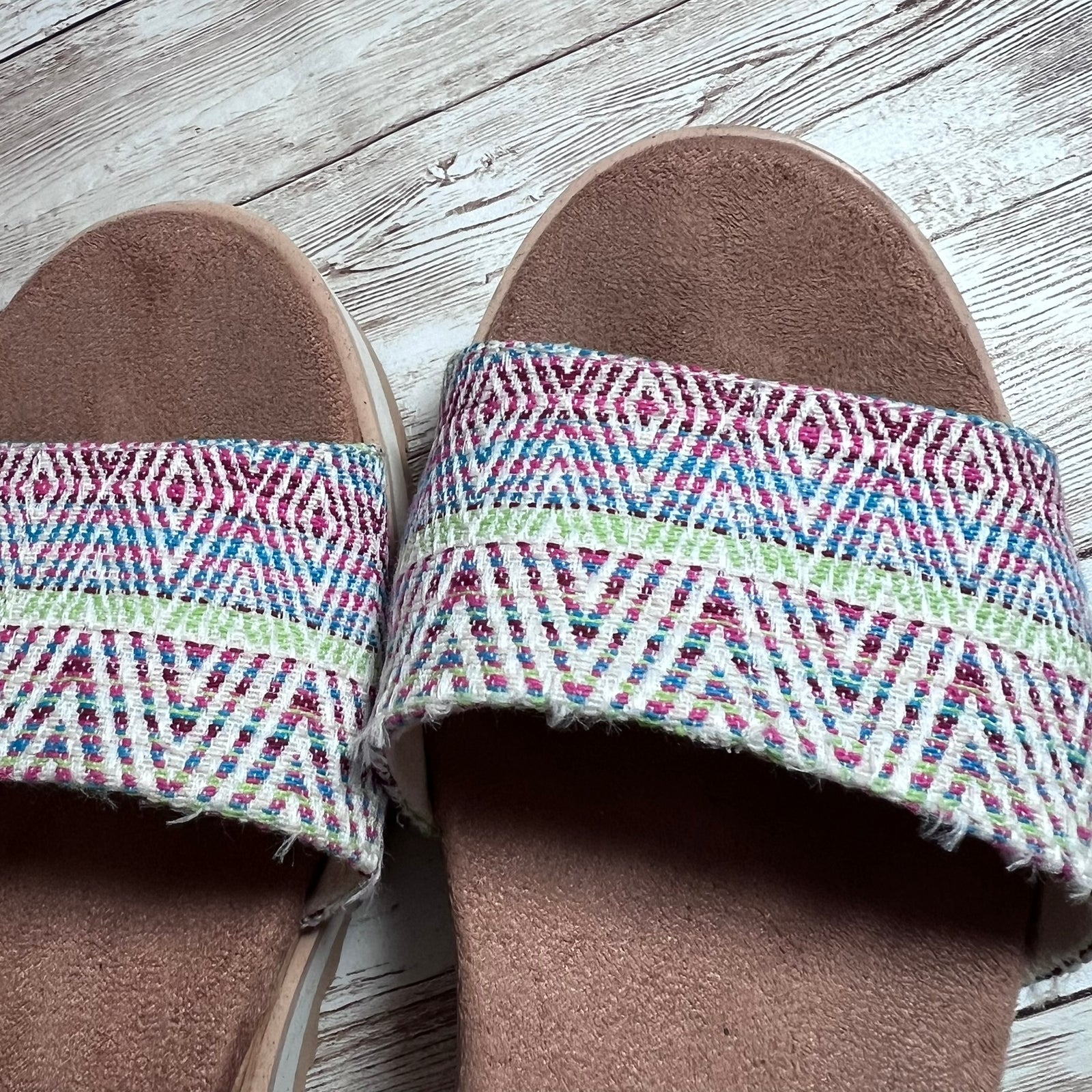 A pair of slide sandals with brown suede-like footbeds and wide woven straps featuring a multicolor geometric pattern in pink, blue, green, and white, shown on a whitewashed wooden floor.