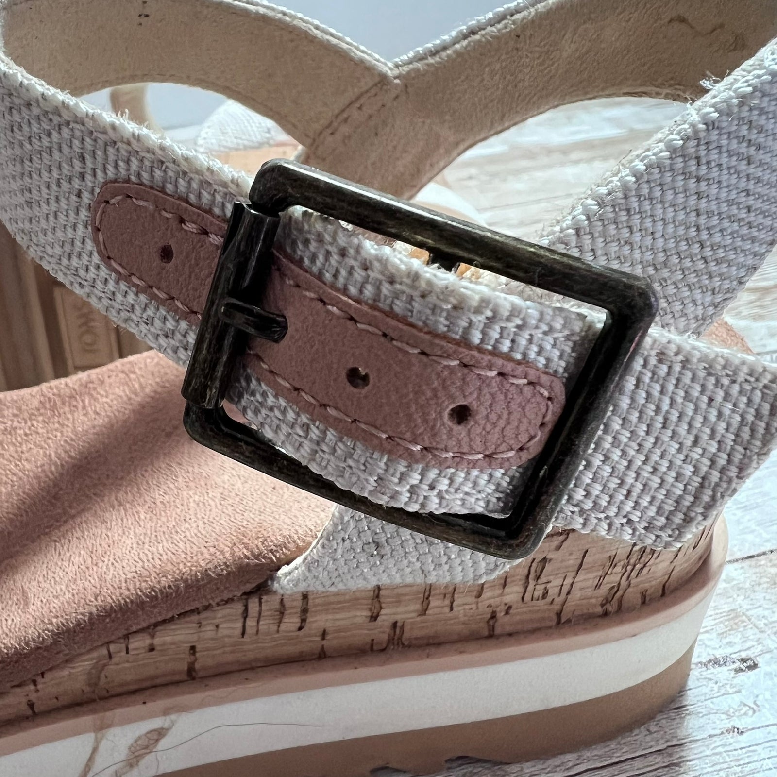 Close-up of a women's wedge sandal with a woven off-white strap, a dusty pink faux-leather inlay, and a black metal buckle, resting on a cork-wrapped wedge heel.
