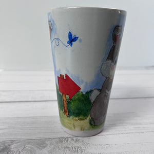 Ceramic mug with a painted design of a red mailbox, blue butterfly, and green field on a white wooden surface.