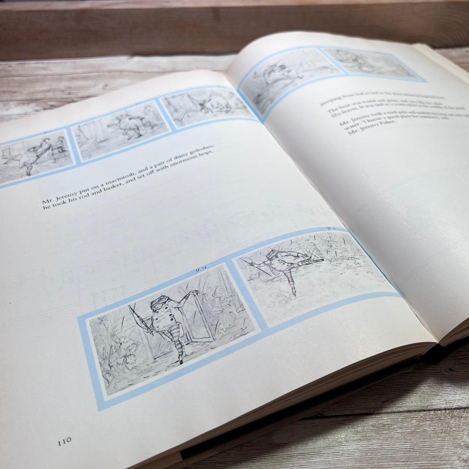An open book lying on a rustic wooden surface, showing printed pages with black line drawings and text framed by pale blue borders.