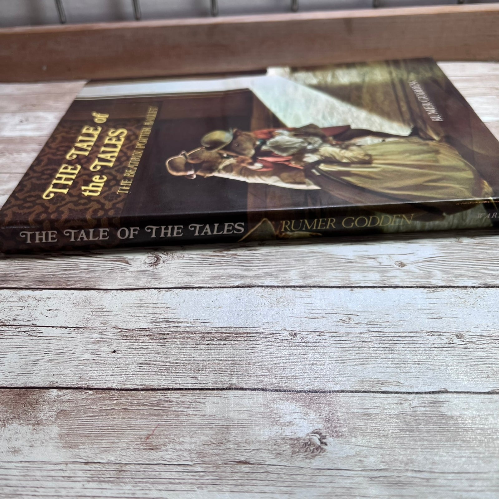 A hardcover book titled "The Tale of the Tales" by Rumer Godden, lying on a rustic light gray wooden surface. The cover features a dark illustration of a knight in armor.
