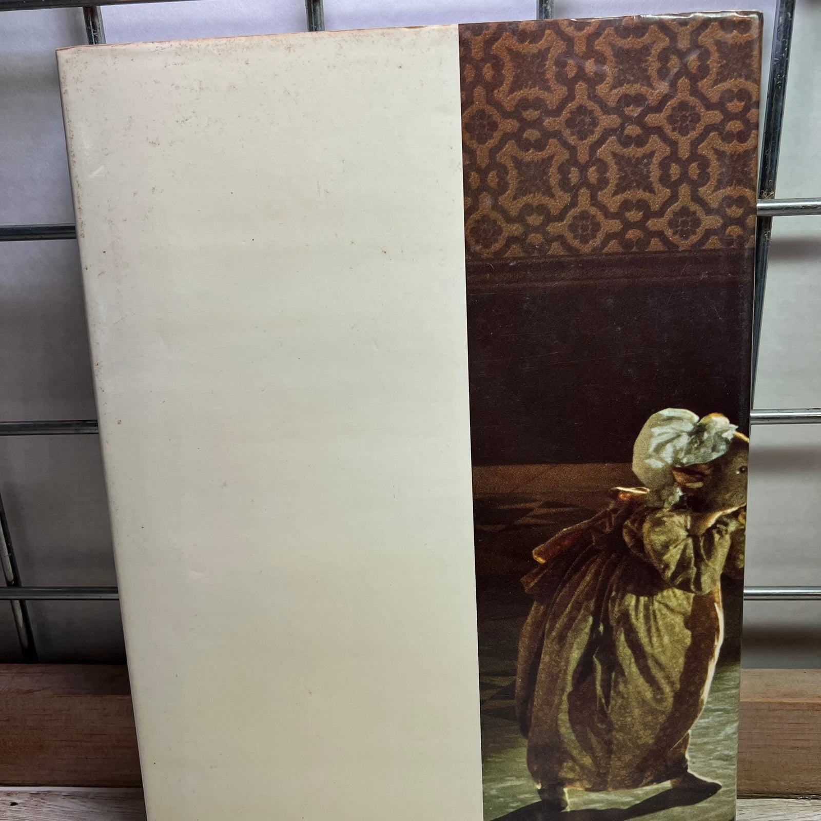 A vintage art print or book cover standing upright on a shelf. The left half is a blank warm off-white/cream area, while the right half shows a classical painting of a woman in a long dress carrying a child, with a brown patterned top section above.