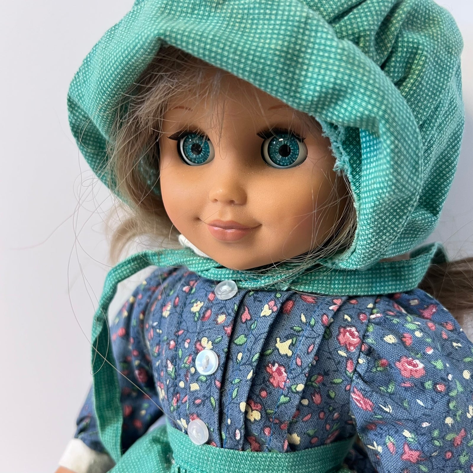 A close-up of a doll with large blue eyes wearing a teal bonnet and a blue floral dress with white buttons, photographed against a plain light background.