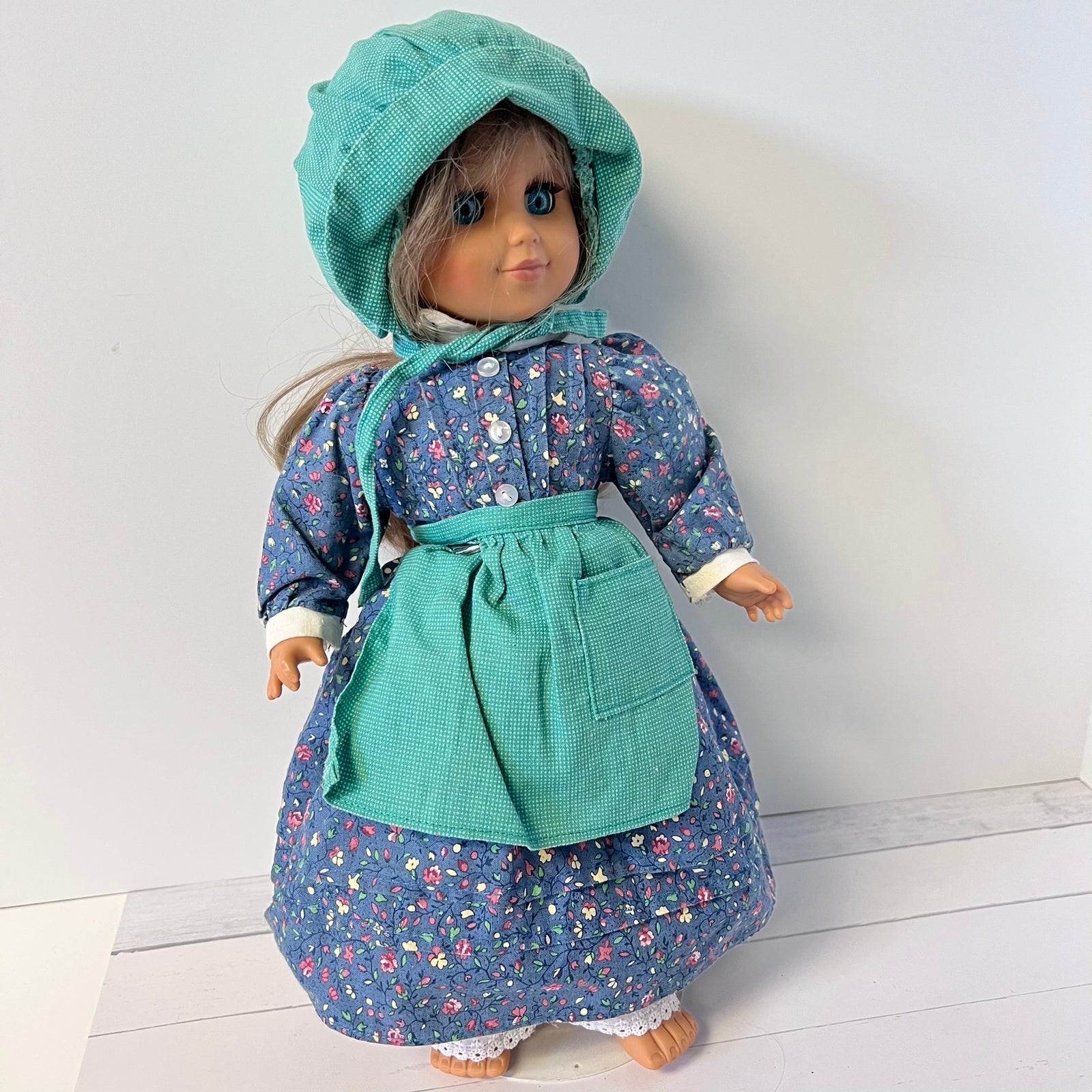 A doll dressed in a floral blue dress with a matching teal apron and bonnet, standing against a plain light gray wall.