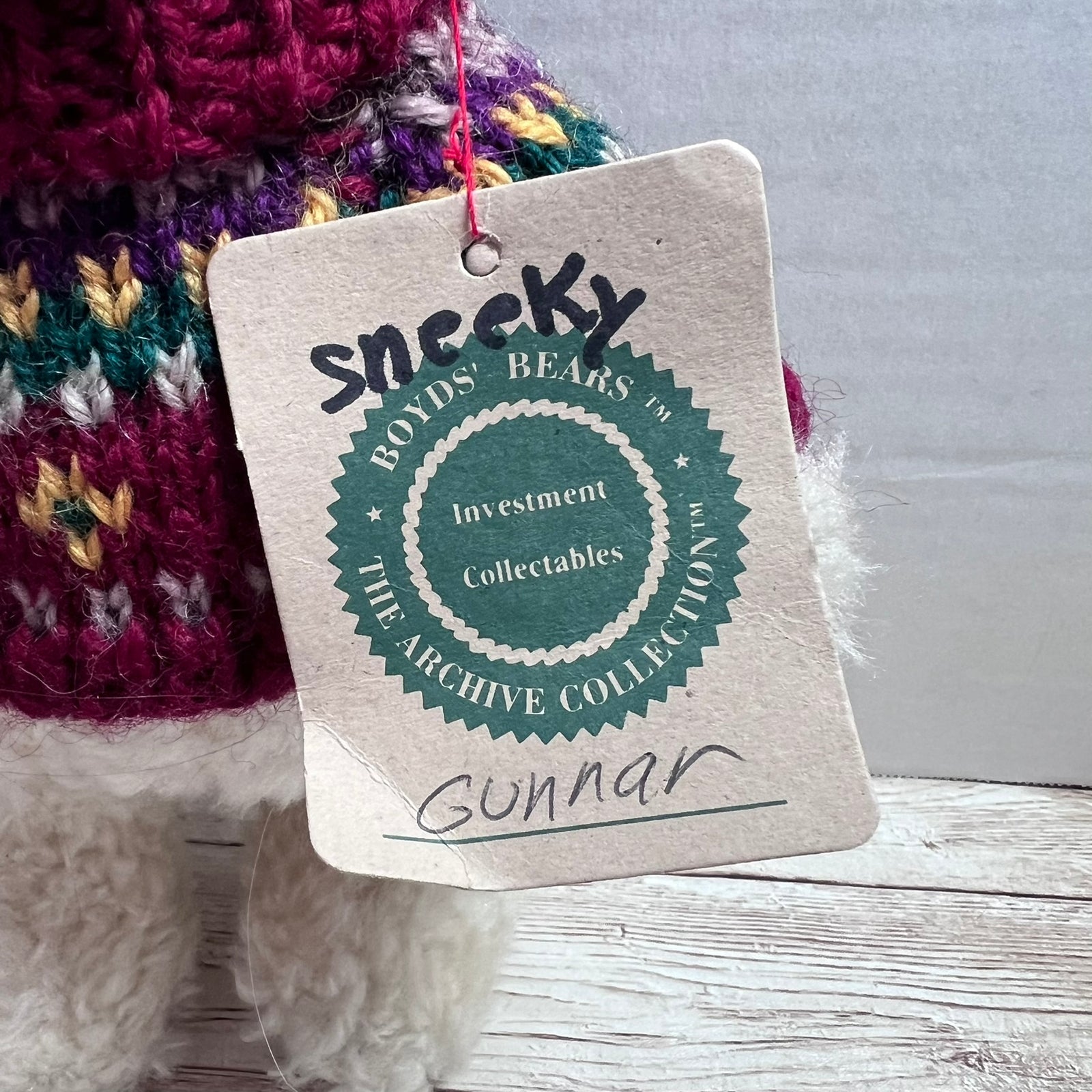 A close-up of a handmade plush toy with a colorful knit hat and a paper hangtag. The tag reads 'Sneeky' and 'Gunnar' with a circular logo for 'Roods' Bears'.