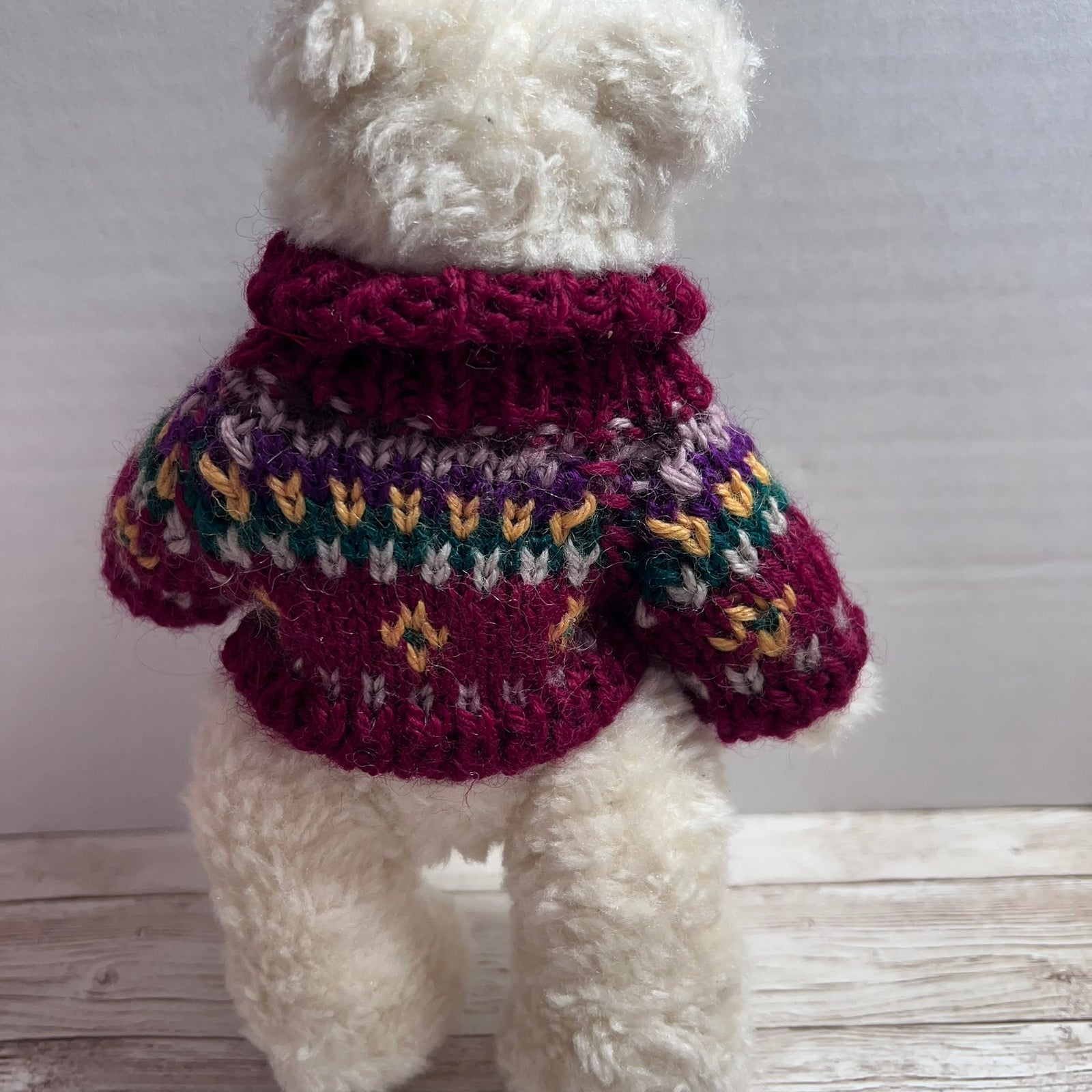 A cream-colored plush teddy bear wearing a hand-knit dog sweater with a burgundy turtleneck and a multicolor Fair Isle pattern on the body.
