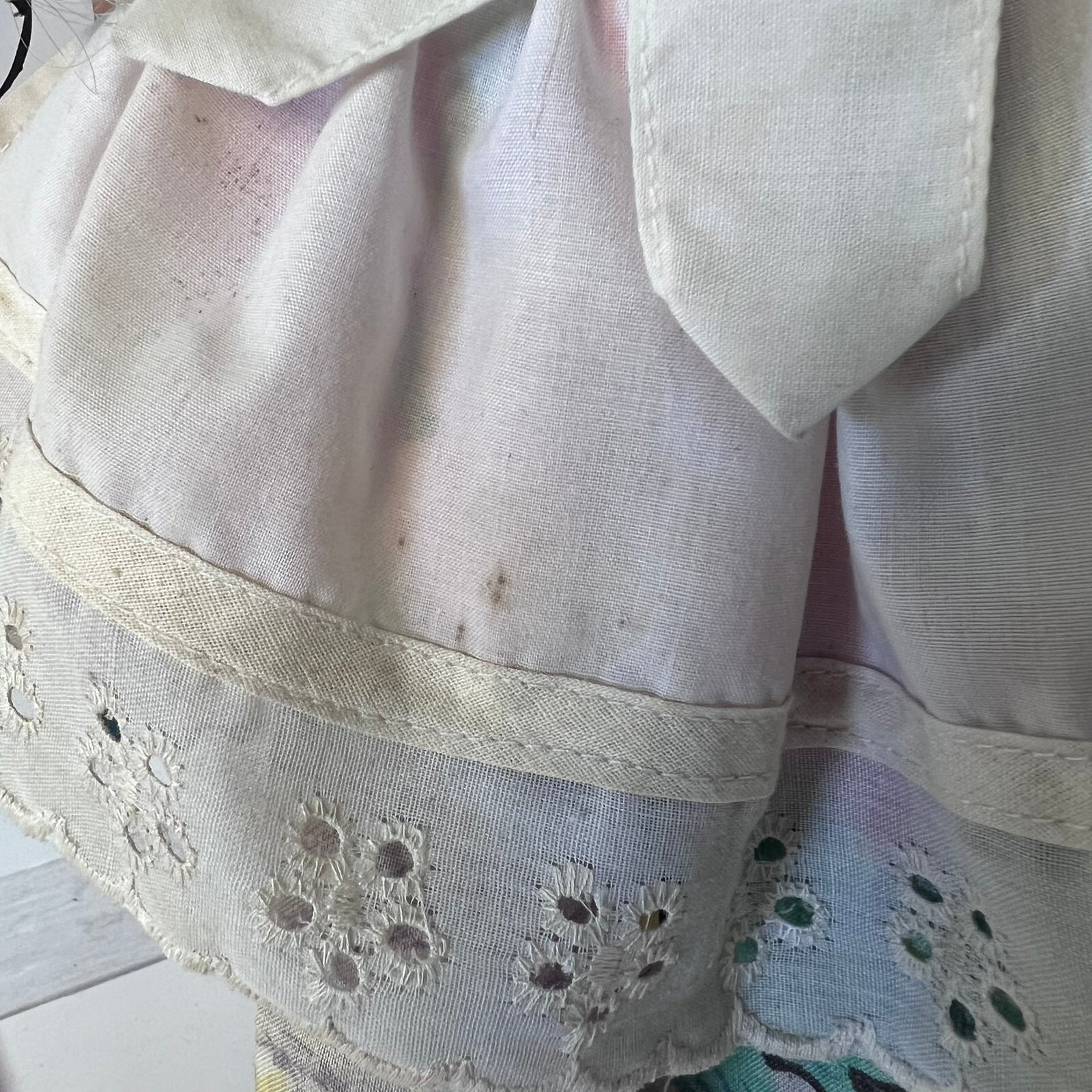 A close-up of a vintage white or off-white cotton dress or skirt fabric, featuring a pointed collar, a decorative band with floral eyelet embroidery, and a soft pastel rainbow sheen over the material.